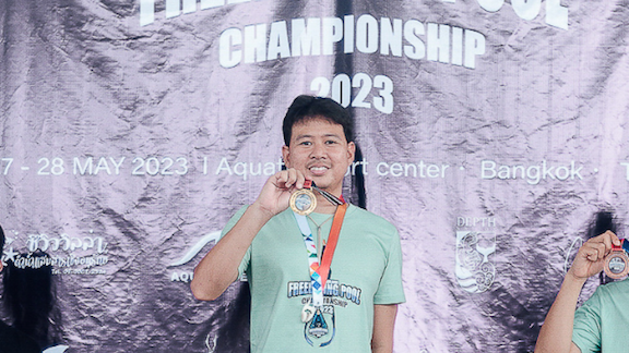 A person stands on a podium at the AIDA Thailand Freediving Pool Championship 2023, smiling and holding a gold medal. They are wearing a light green t-shirt with event logos and text. The background is a crinkled banner with event details, including the date and location in Bangkok. The left side of the fraome is slightly obscured by another individual's arm, also holding a medal.